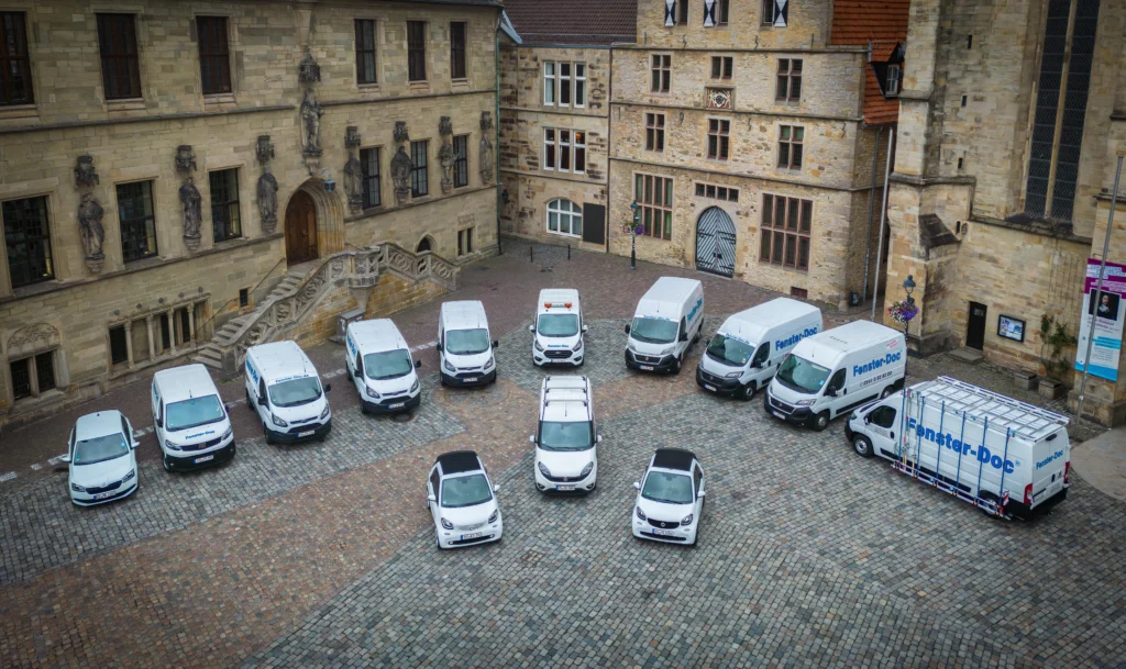 Ein gepflasterter Innenhof mit historischen Gebäuden, umgeben von geparkten „Fenster-Doc“-Fahrzeugen.