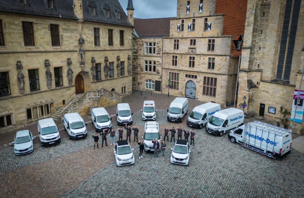 Gruppe von Menschen posiert vor weißen "Fenster-Doc" Lieferwagen, umgeben von historischen Gebäuden.